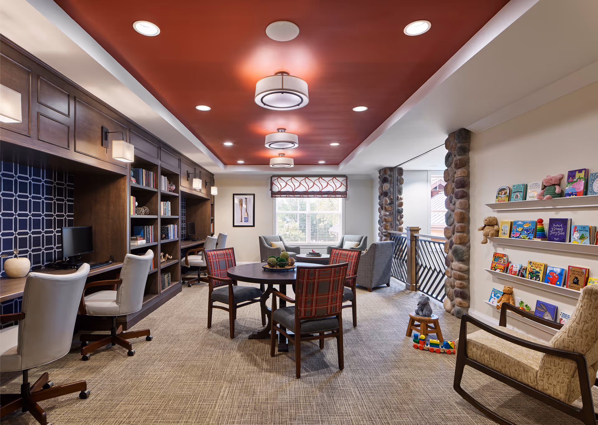 A cozy and well-lit common area with a red ceiling featuring modern light fixtures. The room has a round table with four chairs in the center, a built-in wooden bookshelf and desk area with two white swivel chairs on the left, and a seating area with two armchairs near a window at the back. On the right side, there are shelves with children's books and toys, and a beige armchair in the foreground. Stone pillars and a railing are visible near the window.