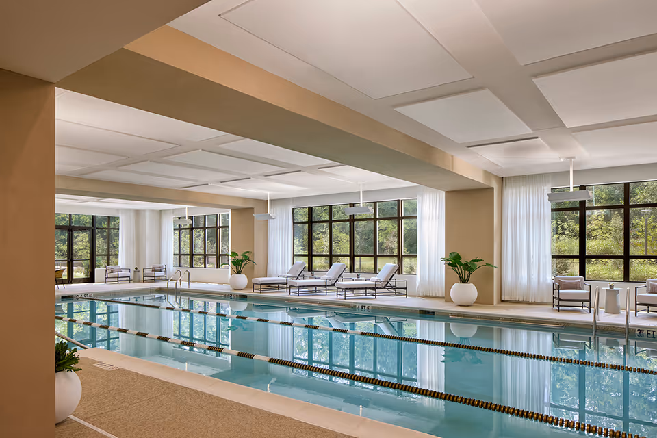 Indoor swimming pool with clear water, lane dividers, and poolside lounge chairs. Large windows with white curtains allow natural light to fill the space, and there are potted plants placed around the pool area.