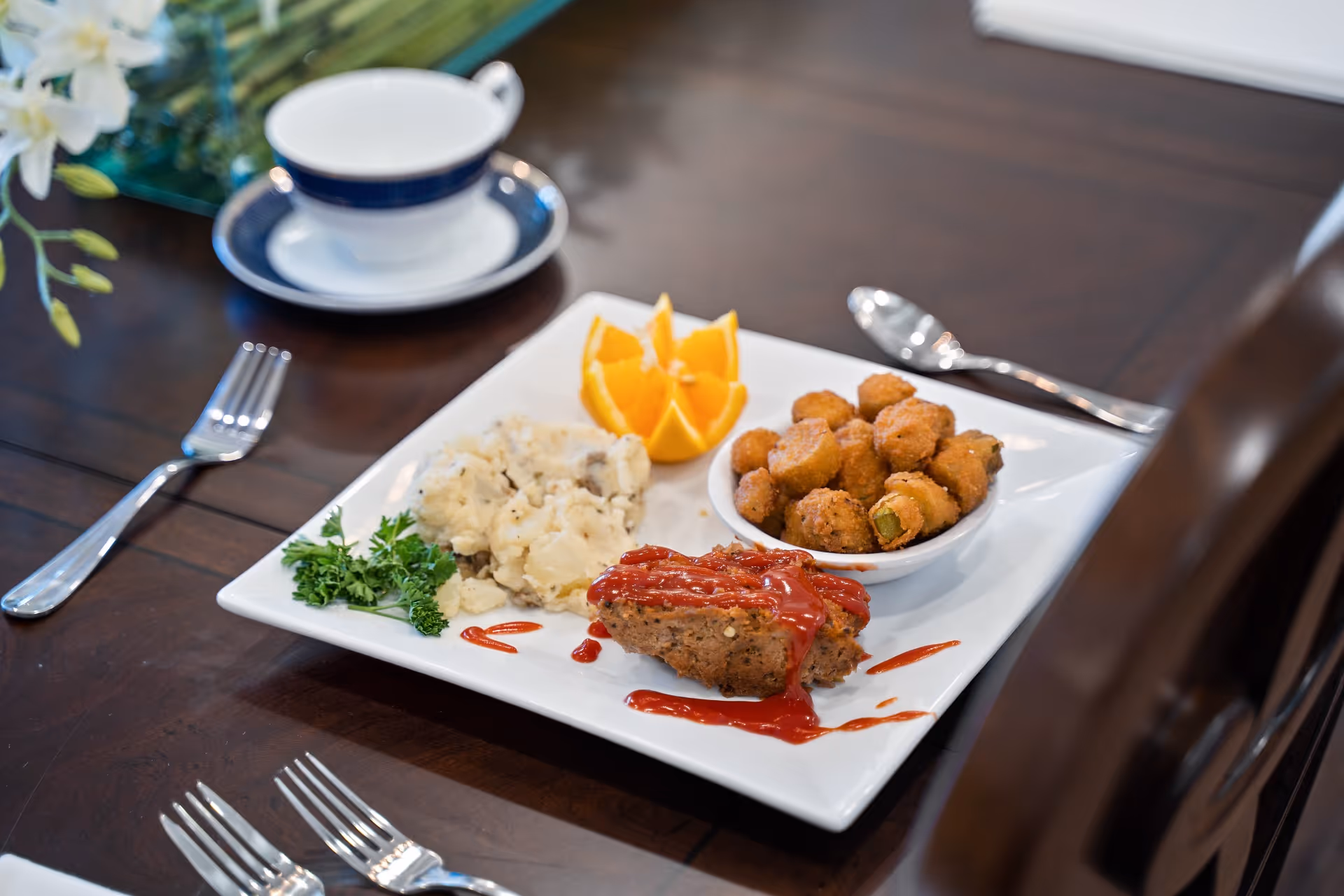 A plated meal on a wooden dining table with meatloaf topped with ketchup, mashed potatoes, fried bites, orange slices, and a teacup.