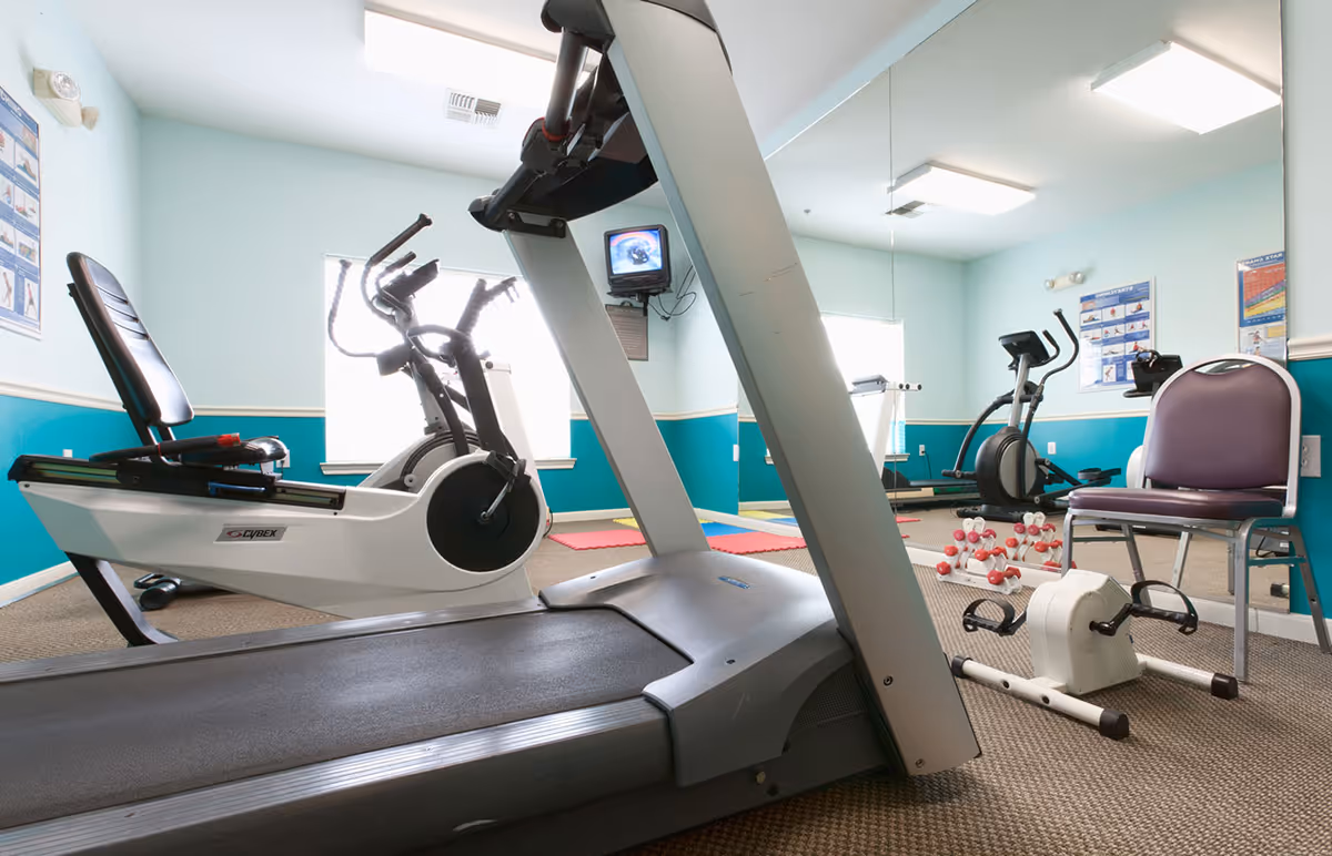 A small fitness room with exercise equipment including a treadmill, stationary bike, elliptical machine, hand weights, and a chair. The room has light blue walls with a large mirror on one side and two windows letting in natural light.