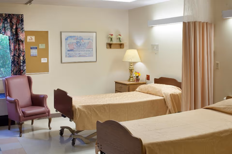 Shared senior care bedroom with two adjustable beds, a bedside table and lamp, a privacy curtain, and a pink armchair by the window.