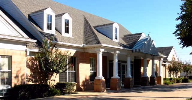 Exterior view of a single-story brick building with white columns supporting a covered entrance. The building has a gray shingled roof with dormer windows and is surrounded by small trees and shrubs under a clear blue sky.