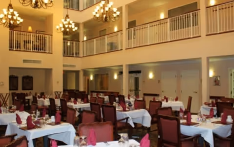 A spacious dining room with multiple tables covered in white tablecloths, each set with glassware and folded red napkins. The room features high ceilings with chandeliers and a second-floor balcony overlooking the area.