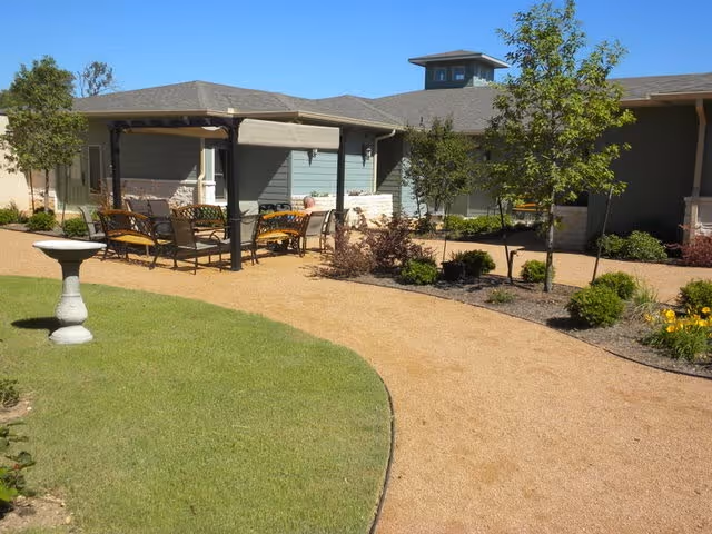 Outdoor patio area at Provident at Buda featuring a gravel pathway, green lawn, a birdbath, and a shaded seating area with chairs and tables surrounded by small trees and shrubs under a clear blue sky.