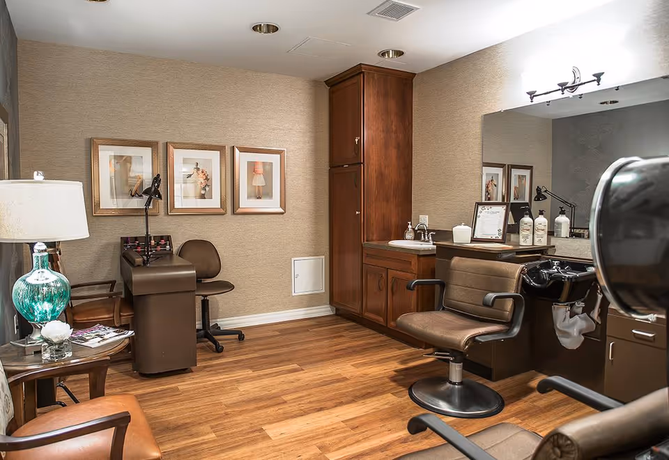 Interior of a salon room with wooden flooring, a styling chair in front of a sink for washing hair, a large mirror, wooden cabinets, a small desk with a chair, and framed pictures on the wall. There is also a side table with a green lamp and magazines.