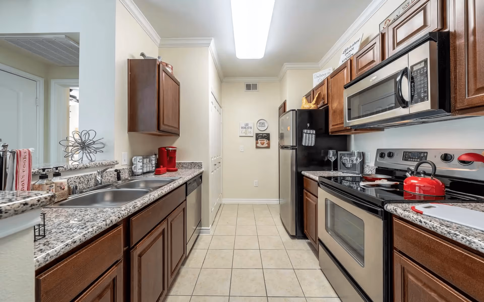 A modern kitchen with granite countertops, wooden cabinets, stainless steel appliances including a microwave, stove, and refrigerator. The kitchen has a double sink, a red coffee maker, and various kitchen accessories. The floor is tiled, and there are decorative signs on the far wall.