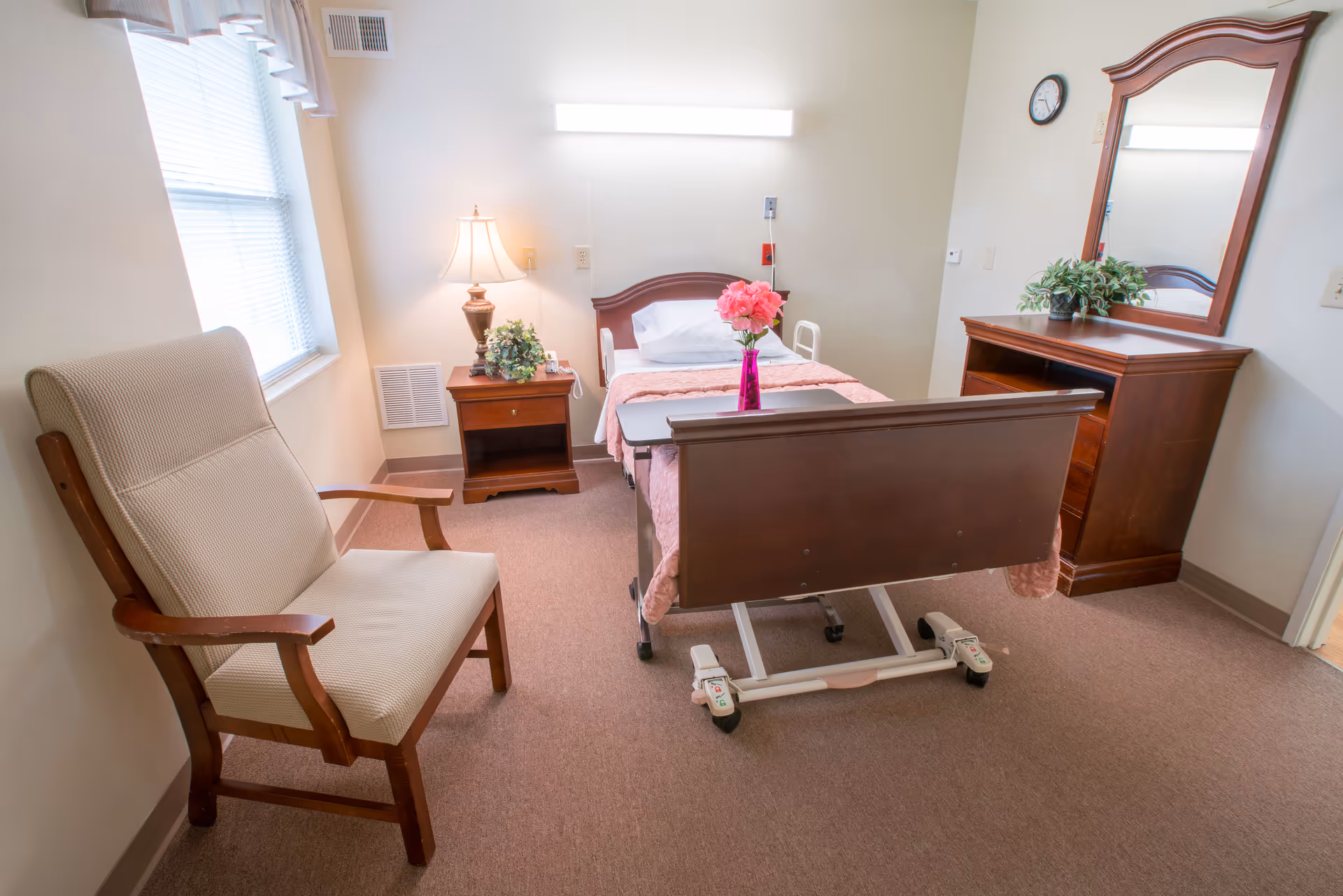 A nursing center bedroom with a single hospital-style bed covered with a pink blanket and a vase of pink flowers on the footboard. There is a wooden nightstand with a lamp and a plant, a cushioned armchair near a window with blinds, and a wooden dresser with a mirror and another plant. The room has beige walls and carpeted floor.