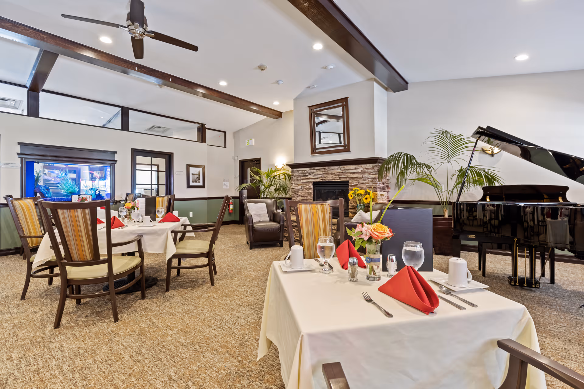 A well-lit dining area in a senior living facility with tables set with white tablecloths, red napkins, glassware, and floral centerpieces. The room features a stone fireplace with a mirror above it, a grand piano, comfortable chairs, and ceiling fans. There are plants and framed pictures decorating the space.