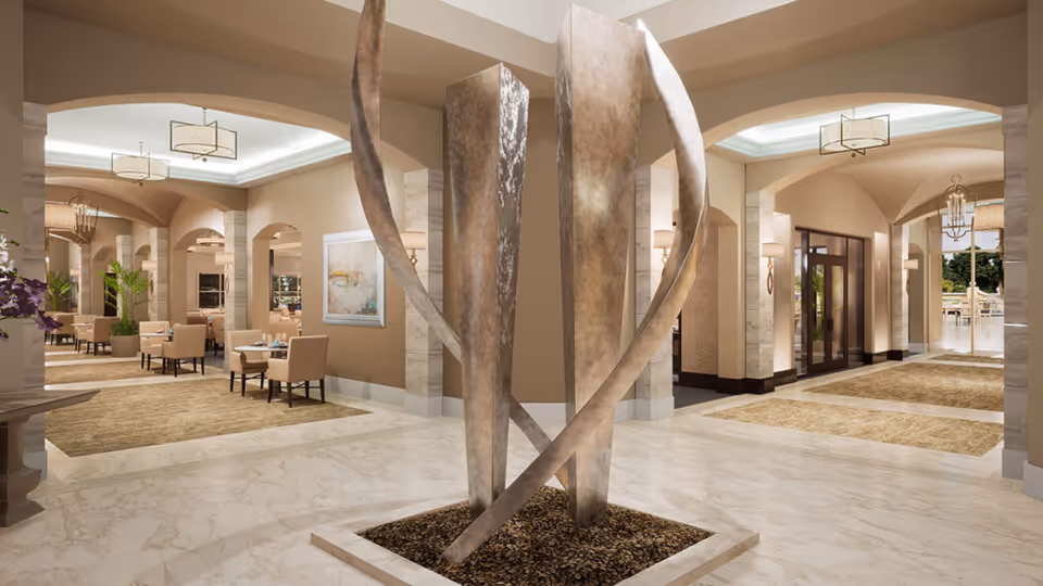 A spacious and elegant interior common area with a modern abstract metal sculpture in the center. The room features marble flooring, beige walls, and arched doorways leading to seating areas with tables and chairs. Soft lighting fixtures hang from the ceiling, and large windows provide natural light.