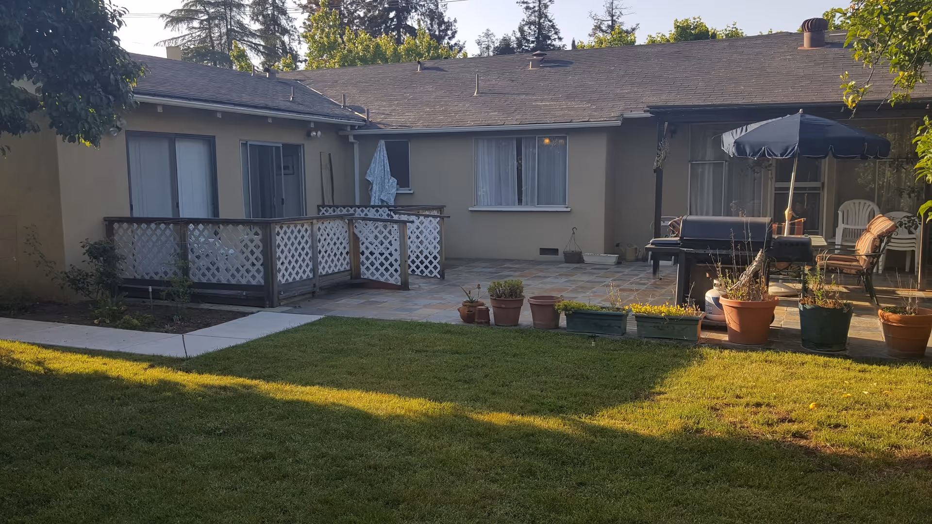 Backyard view of a single-story residential building with a small patio area. The patio has several potted plants, a barbecue grill, and outdoor seating with an umbrella. There is a grassy lawn in the foreground and some trees and shrubs around the edges.
