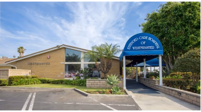 Exterior view of Extended Care Hospital of Westminster showing a single-story brick building with large windows, a blue awning entrance with the facility name, palm trees, and landscaped greenery under a partly cloudy sky.