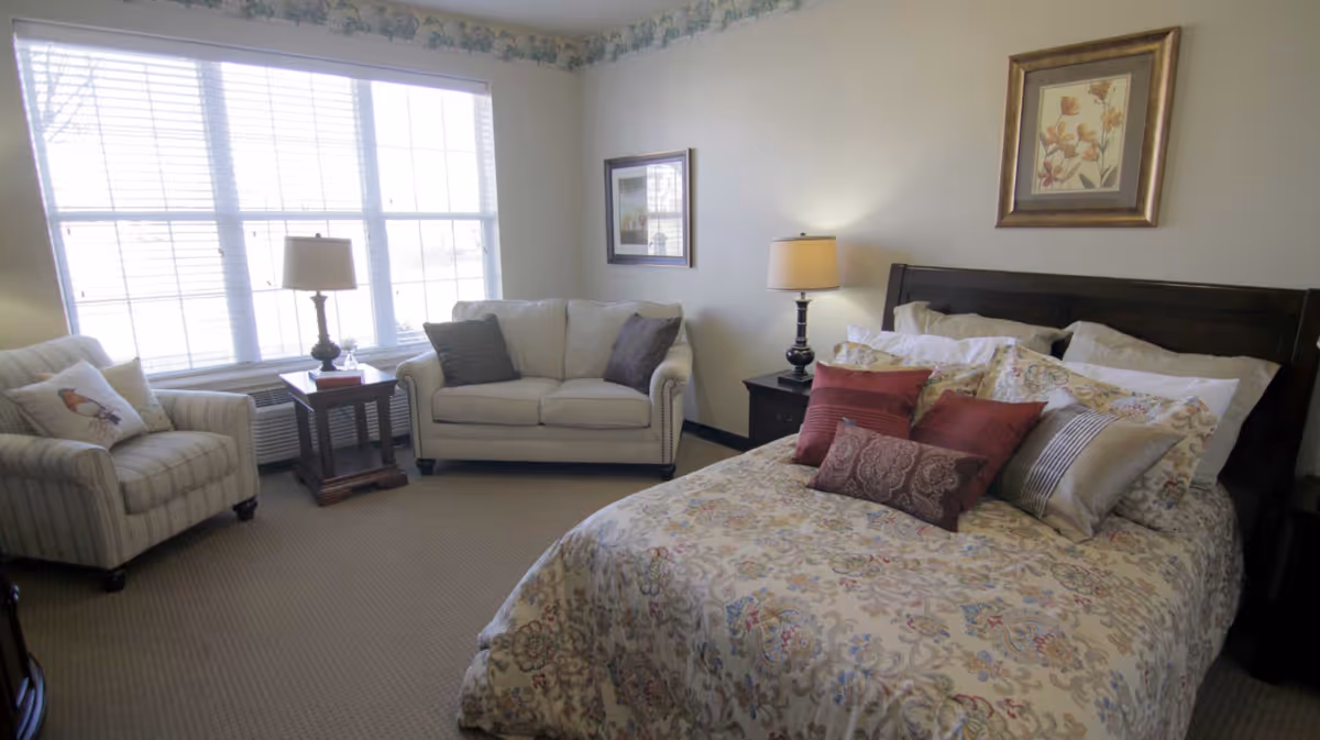 Furnished bedroom with a floral-patterned bed, decorative pillows, nightstands with lamps, and a seating area by a large window.