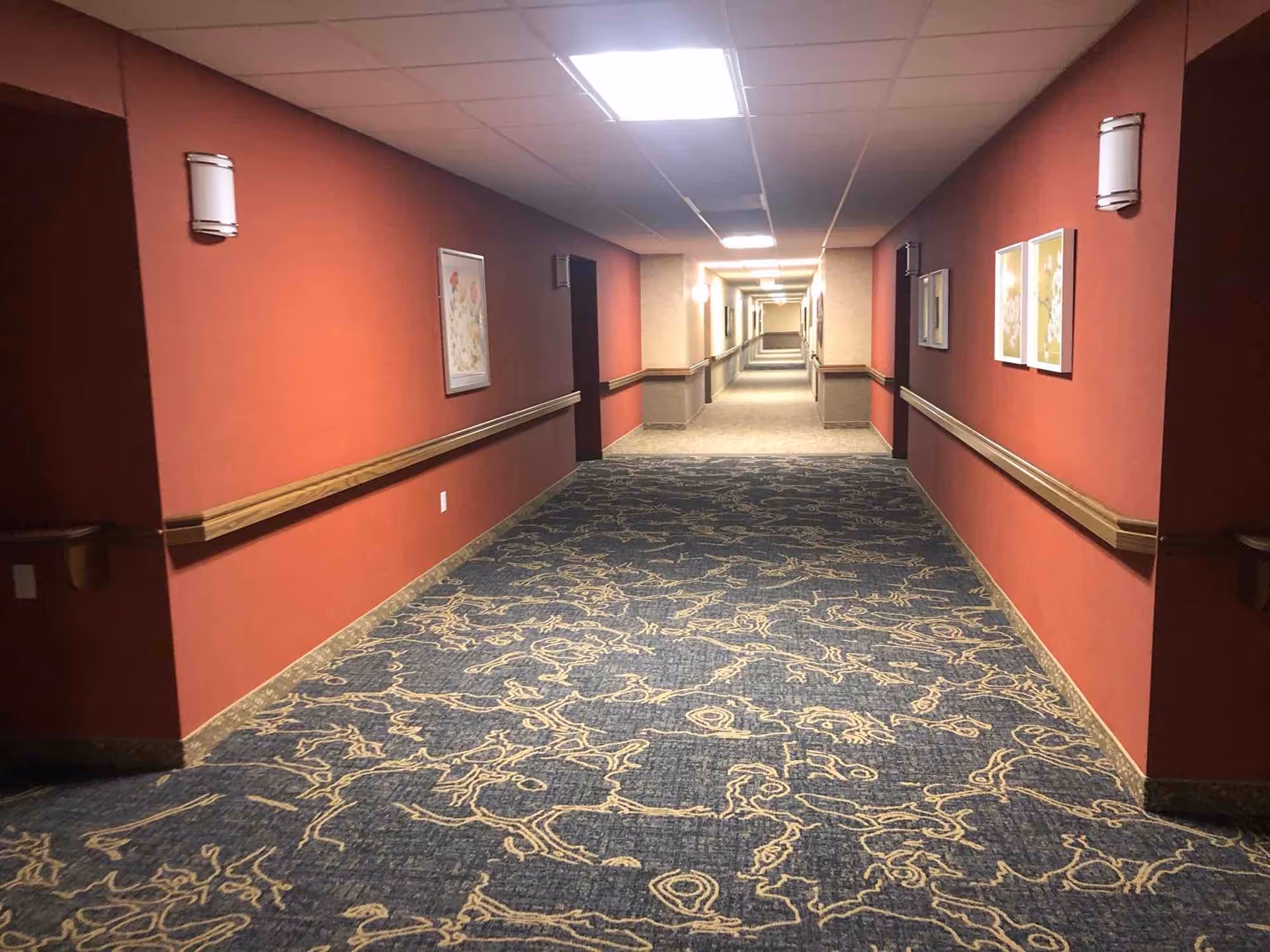Long carpeted interior hallway in a retirement community with red walls, handrails, ceiling lights, and framed artwork.