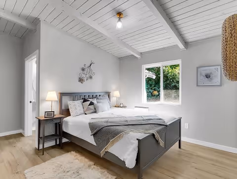 A bright and cozy bedroom with a gray wooden bed frame, white bedding, and decorative pillows. Two bedside tables each hold a lamp, and a small clock is on the left table. The room has light gray walls, a window showing greenery outside, a fluffy white rug on the wooden floor, and a decorative wall piece above the bed. The ceiling features exposed beams and a single light bulb fixture.