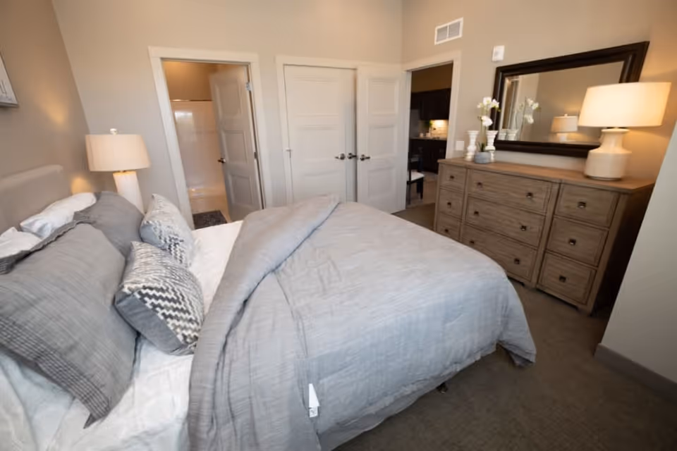 A cozy bedroom in a senior living apartment featuring a neatly made bed with gray bedding and multiple pillows. To the right, there is a wooden dresser with a large mirror above it and a lamp on top. The room has beige walls and carpeted floors. Two doors are visible, one leading to a bathroom and the other to another room, possibly a kitchen.
