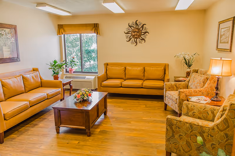 A cozy living room with two tan leather sofas, two patterned armchairs, a wooden coffee table with a floral centerpiece, a side table with a lamp and flowers, a window with a yellow valance, and wall decorations including a sun sculpture and framed pictures.