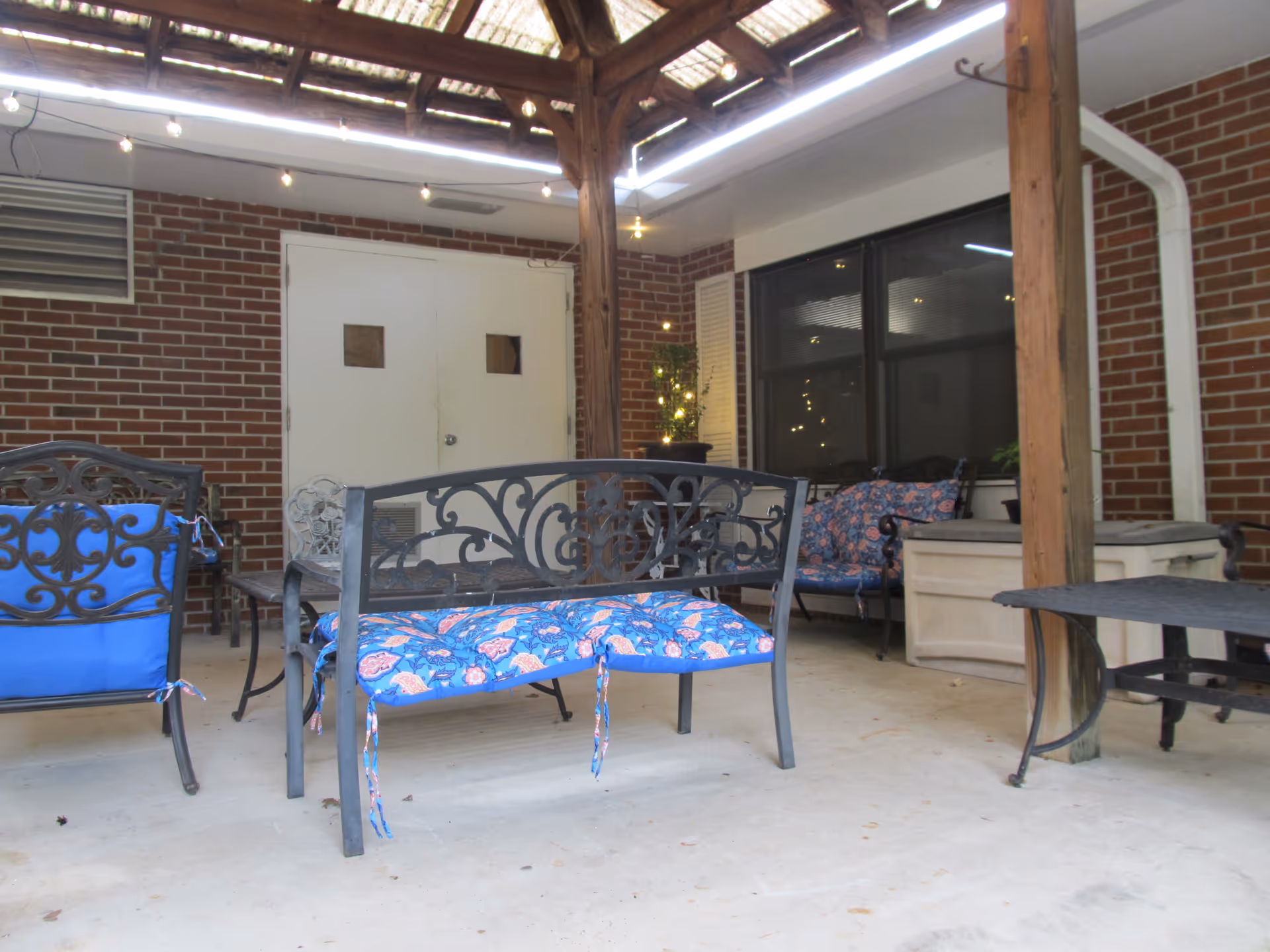 Covered outdoor patio area with metal benches and chairs that have blue and floral patterned cushions. The patio has a wooden pergola roof with string lights hanging from it. There is a brick wall with a white double door and a large window with black frames. A potted plant with lights is visible near the window.