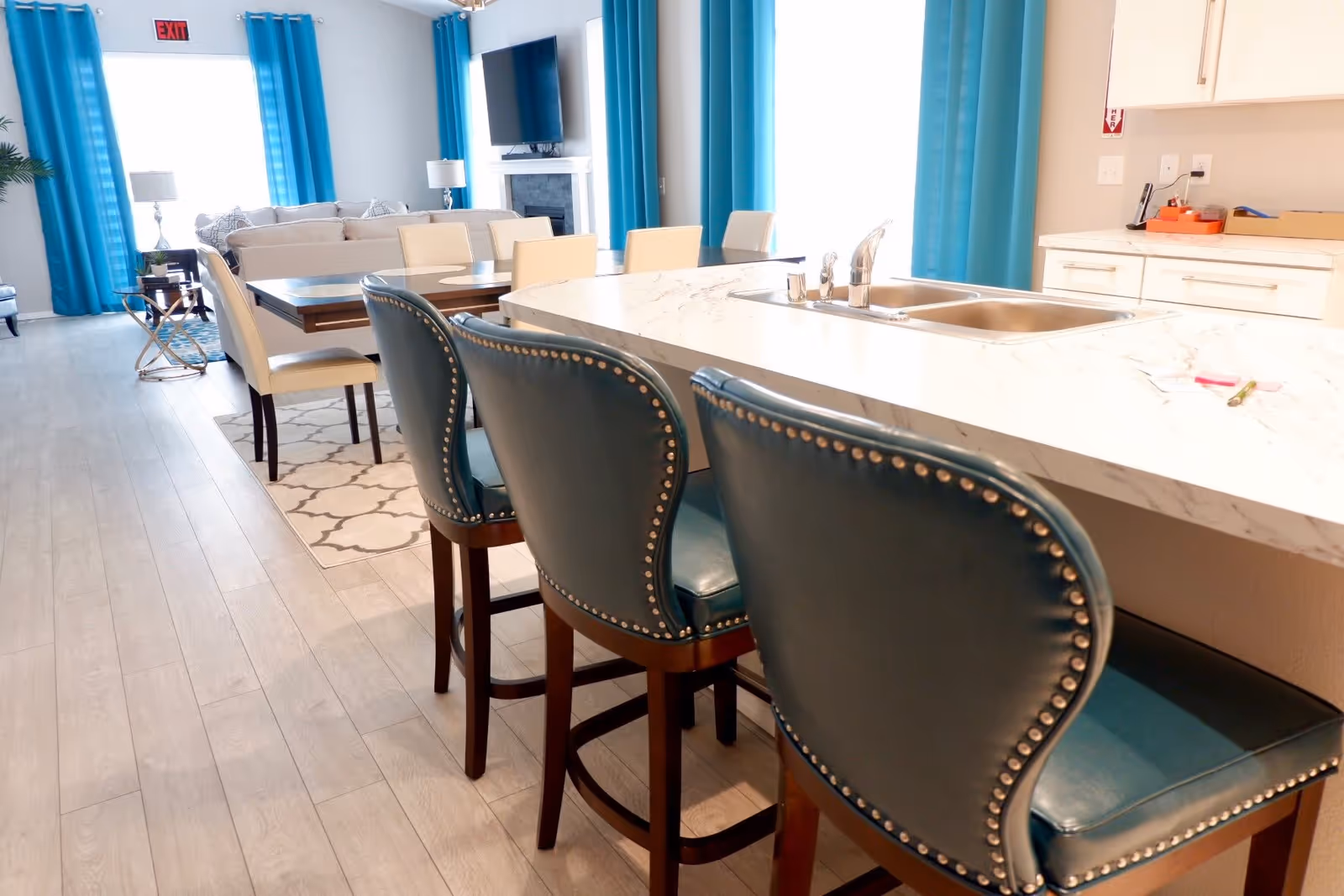 Interior view of a bright and modern assisted living home featuring a kitchen island with three blue cushioned bar stools, a double sink, and white marble countertop. In the background, there is a dining table with cream-colored chairs, a beige sofa, a TV mounted above a fireplace, and large windows with blue curtains allowing natural light to fill the room.