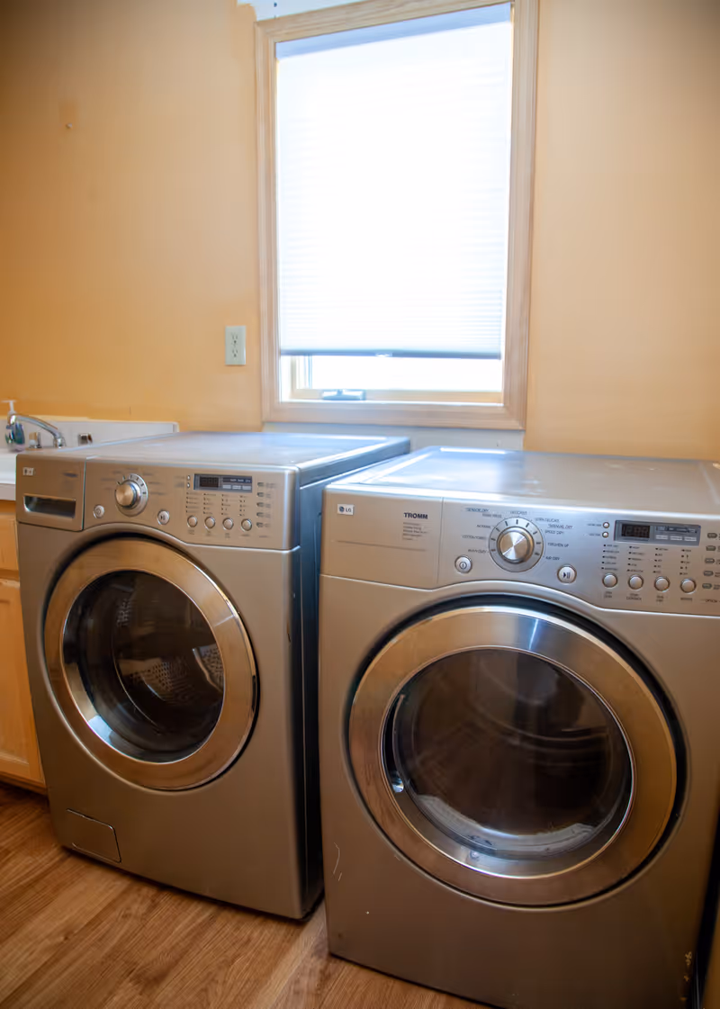 A laundry room with a front-loading washing machine and dryer side by side under a window with a closed blind. The walls are painted a light peach color and the floor has a wood-like finish.