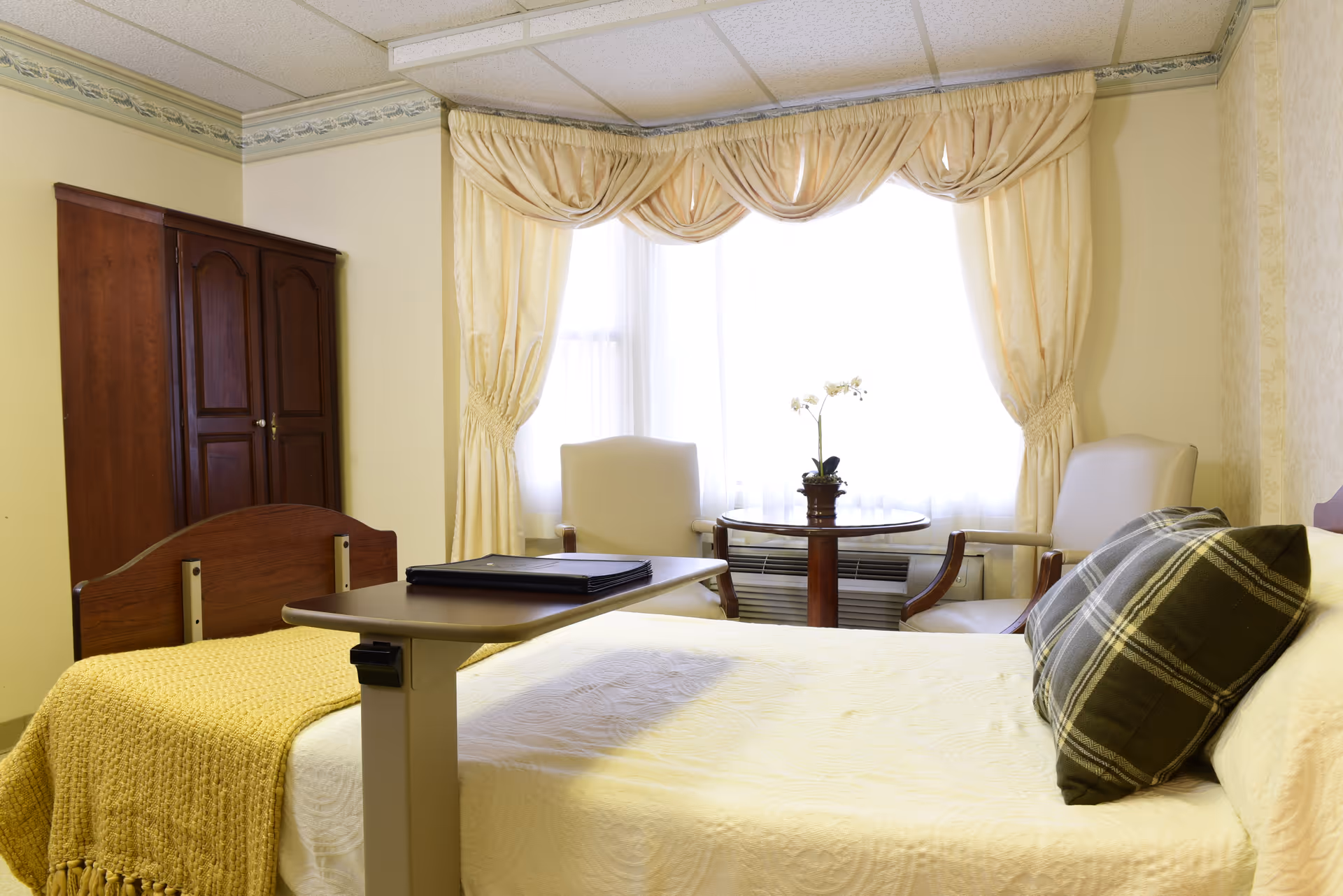 A cozy bedroom in a senior living facility with a single bed covered in a white bedspread and a yellow knitted blanket. There is a plaid pillow on the bed. Next to the bed is an adjustable overbed table with a closed binder on it. In the background, two cream-colored armchairs are positioned around a small round wooden table with a potted orchid. The window behind them is covered with sheer white curtains and beige drapes with a swag valance. A wooden wardrobe is against the wall to the left.