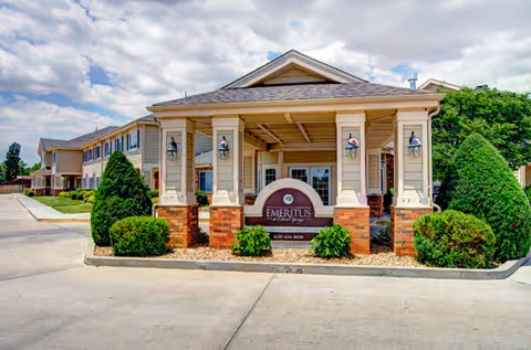 Front exterior view of the Brookdale Liberal Springs facility showing a covered entrance with a sign that reads 'Emeritus at Liberal Springs' surrounded by neatly trimmed bushes and a paved driveway under a partly cloudy sky.