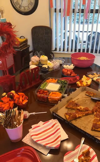 A dining table set with various foods including pizza slices in an open box, a vegetable tray with dip, strawberries, cupcakes, and other snacks. There are utensils in a pink cup, napkins with red and white stripes, and a decorative red 'LOVE' sign on the table. In the background, there is a chair, a clock on the wall, and vertical blinds covering a window.