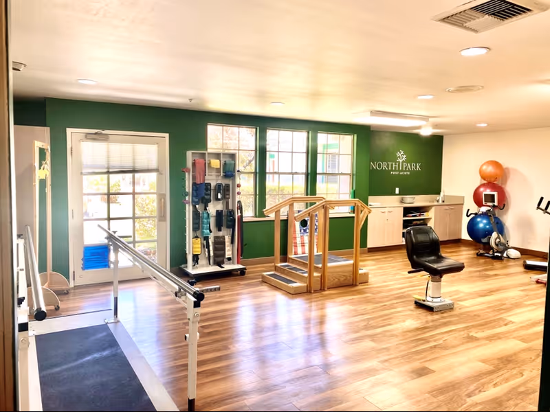 Interior therapy/fitness room with parallel bars, exercise equipment, balance balls, and a green wall reading "North Park Post-Acute".