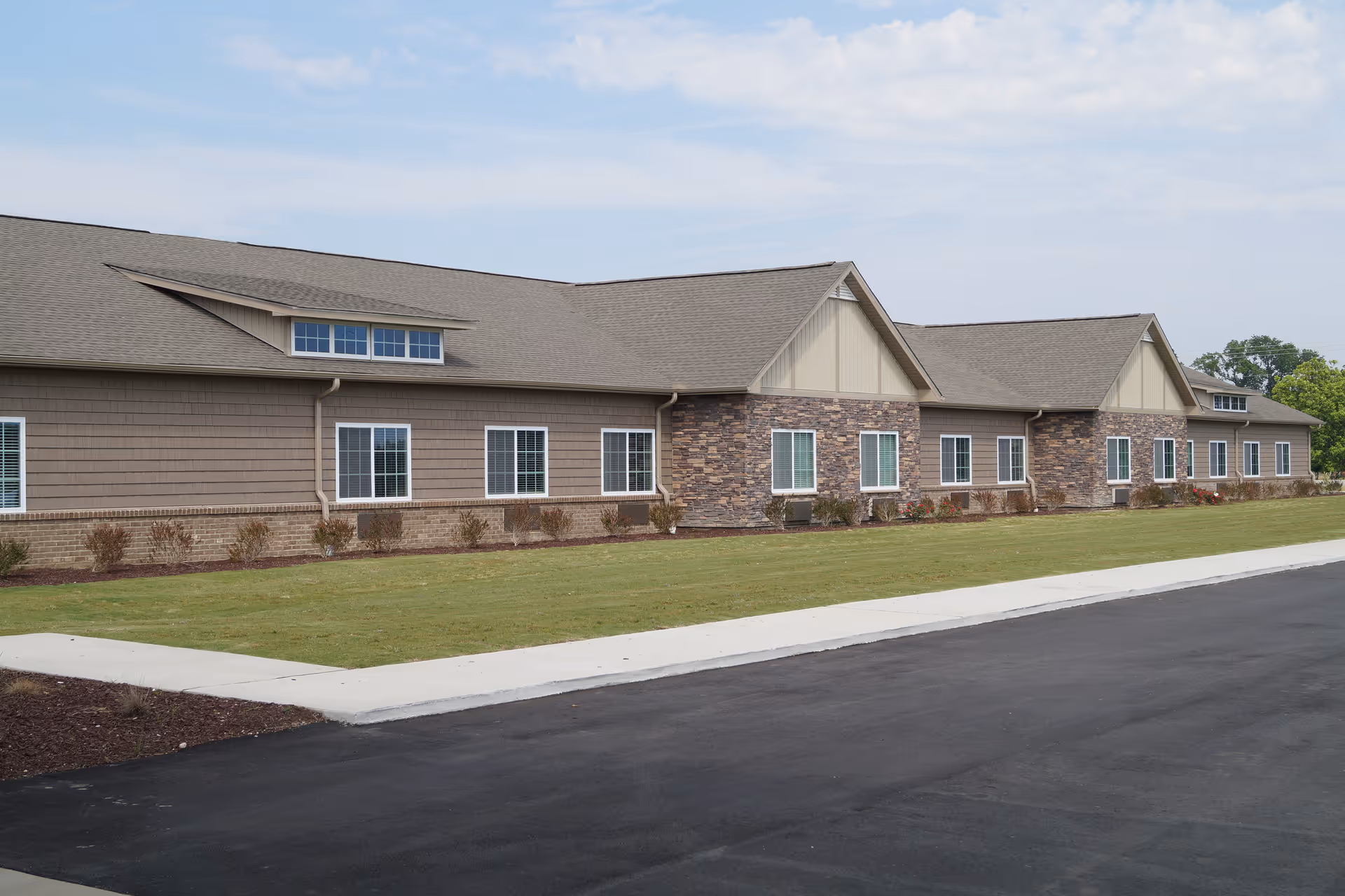 Single-story senior living building with stone and siding facade, multiple windows, a grassy lawn and paved driveway in front.