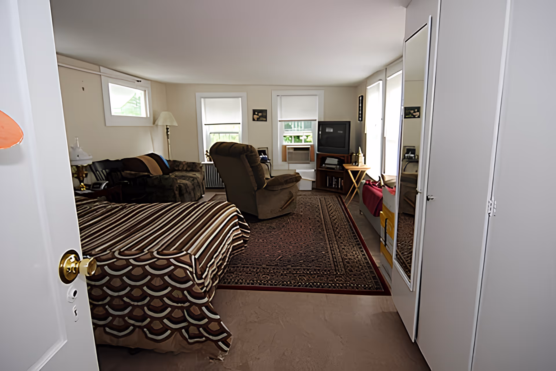A furnished studio-style room with a bed in the foreground, a sofa and recliner facing a television by the windows.