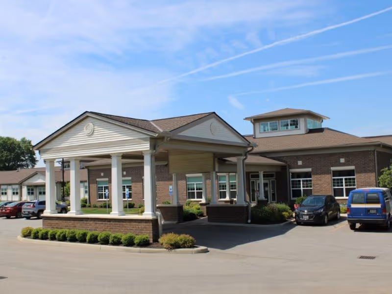 Exterior view of Avenue at Wooster Care and Rehabilitation Center showing a brick building with a covered entrance supported by white columns, several parked cars, and a clear blue sky with some clouds.