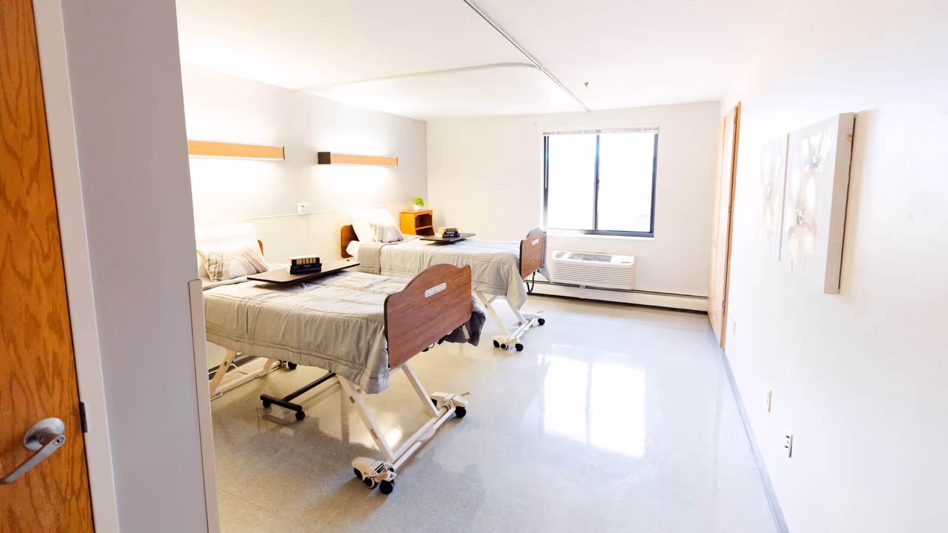 A bright and clean healthcare facility room with two hospital beds, each equipped with side rails and wheels. The beds have light-colored bedding and pillows. There is a window letting in natural light, a wall-mounted air conditioning unit below the window, and two pieces of floral artwork on the right wall. The floor is shiny and reflective, and the room has a simple, uncluttered appearance.