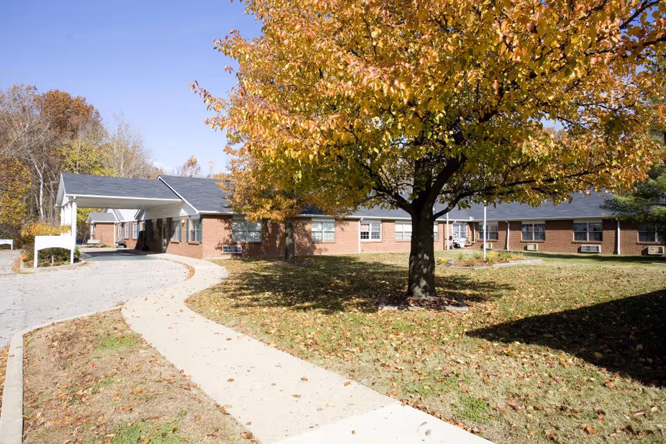 Exterior view of Wedgewood Healthcare Center showing a single-story brick building with a covered entrance. A curved sidewalk leads to the entrance, and a large tree with yellow and orange autumn leaves is in the foreground. The sky is clear and blue.