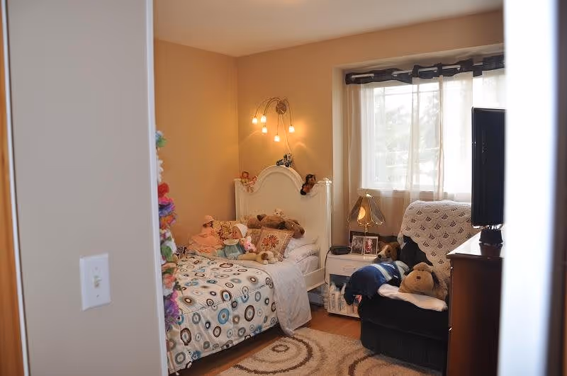 A cozy bedroom with a single bed covered in a patterned bedspread and several stuffed animals and dolls arranged on it. A small nightstand beside the bed holds a lamp and framed photos. Next to the nightstand is a dark armchair with a crocheted blanket draped over the back and more stuffed animals on the seat. A window with sheer curtains allows natural light into the room.