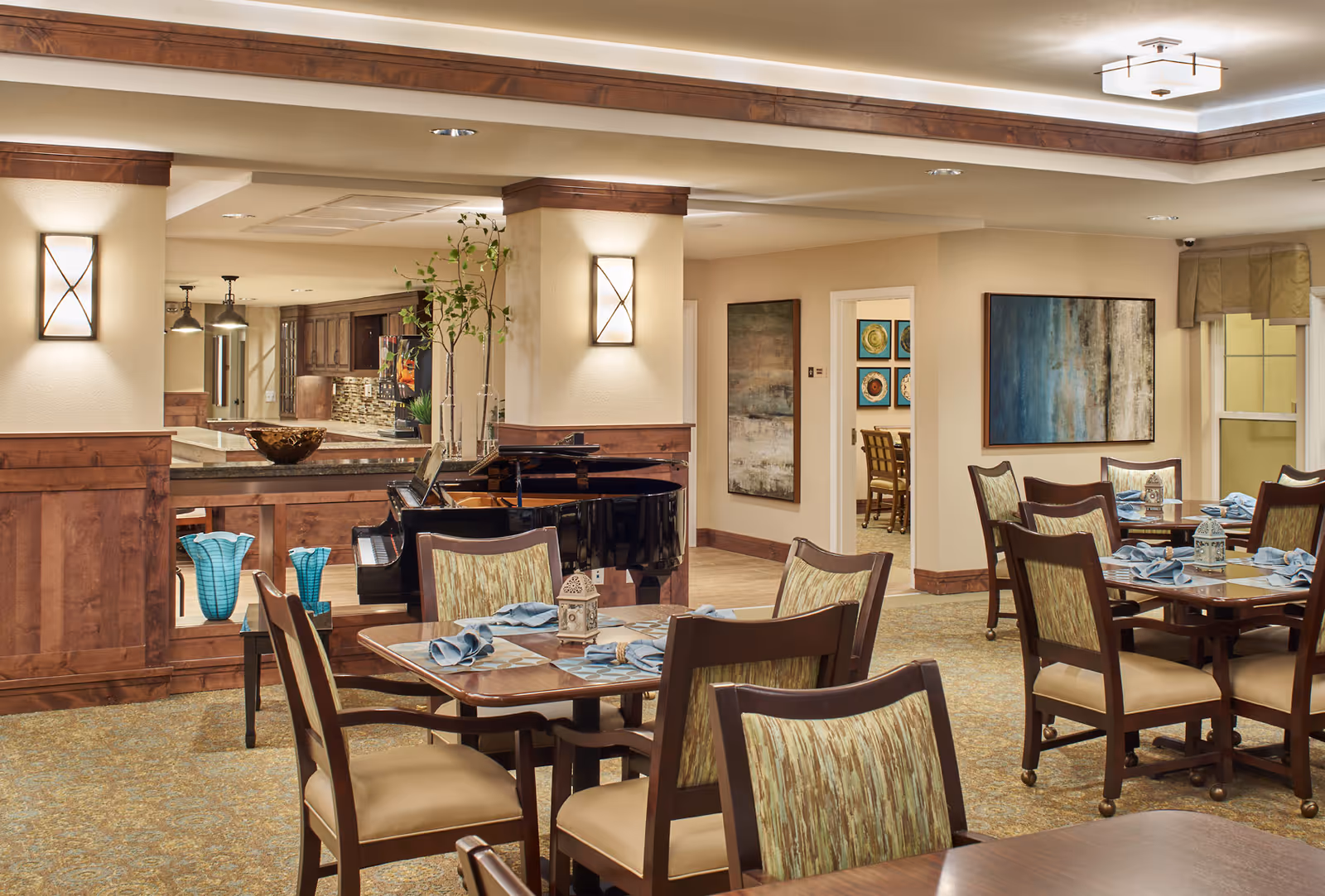 A warmly lit dining room with set tables and chairs, a grand piano, and a service counter opening to a kitchen.