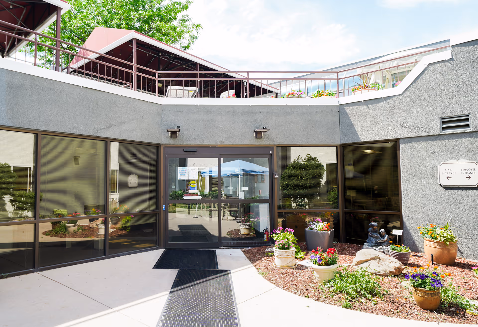 Front entrance of a health care facility with glass double doors, potted flowers, and a walkway leading to the entry.