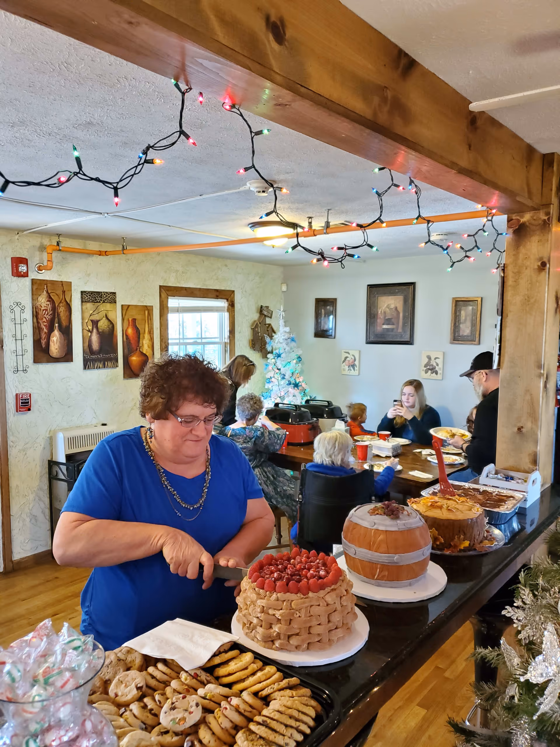 A woman in a blue shirt is cutting a cake decorated with raspberries and a basket-weave frosting pattern on a counter filled with cookies and other cakes. In the background, several people, including elderly individuals and a child, are seated around a dining table in a warmly decorated room with holiday lights hanging from the ceiling and a white Christmas tree.