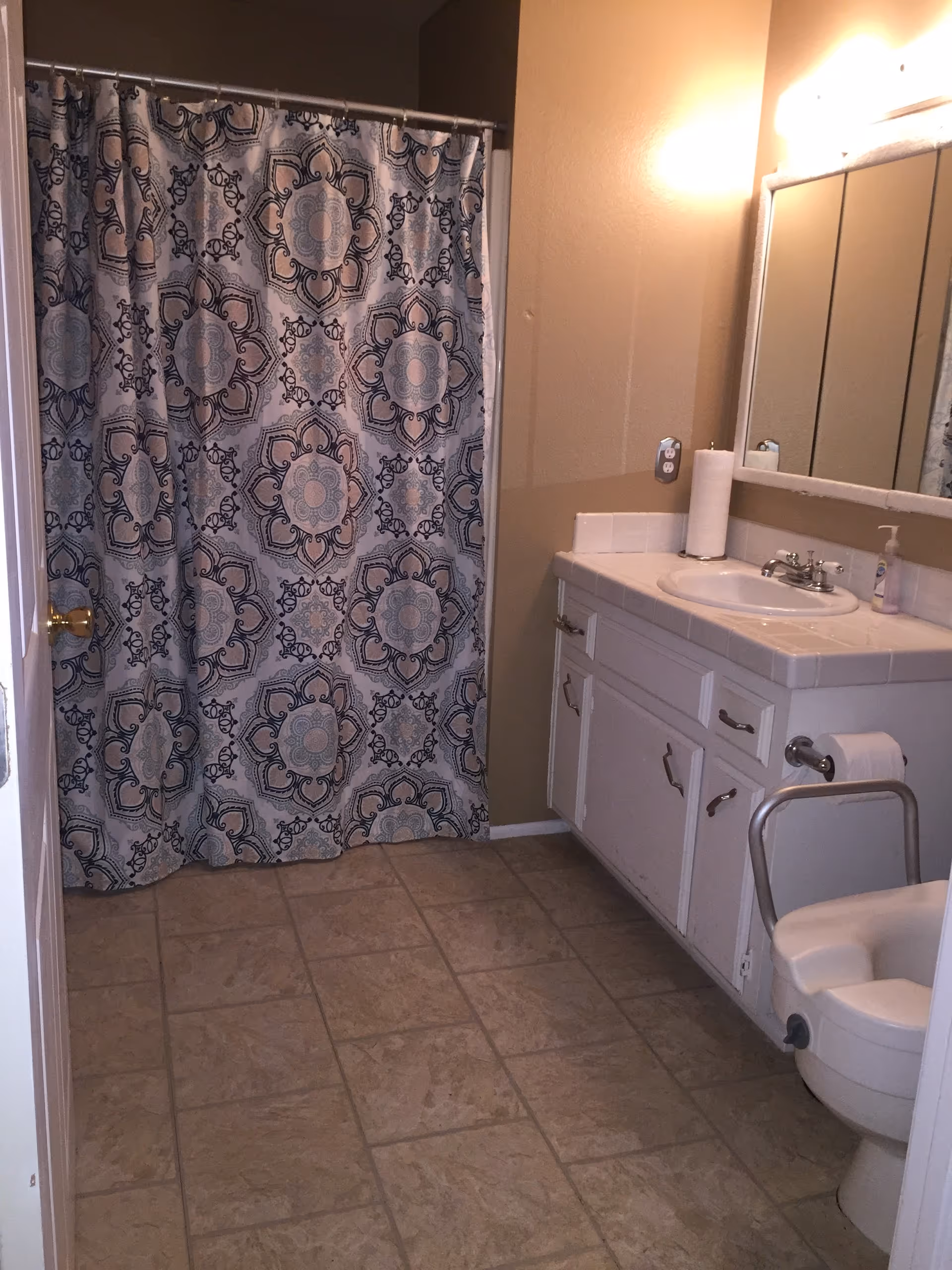 A bathroom with a patterned shower curtain, tiled floor, white vanity with sink and mirror, and a toilet with a safety rail.