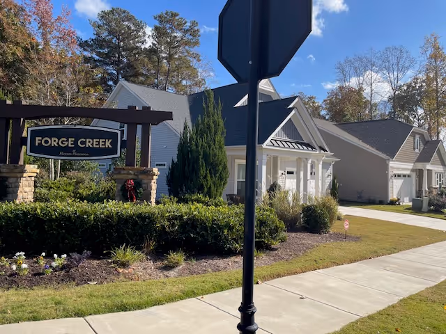 View of the entrance to Forge Creek at Flowers Plantation, showing a landscaped area with bushes and flowers, a sign with the facility name, a sidewalk, and residential-style buildings under a blue sky with some clouds.