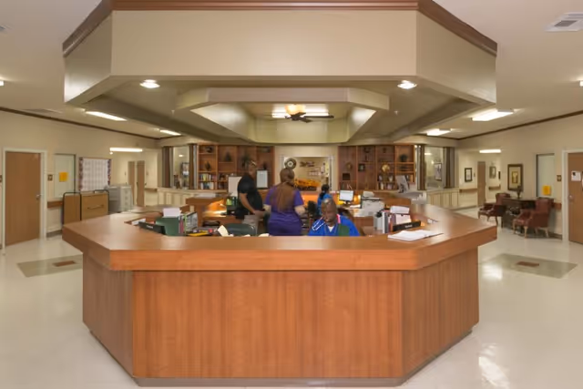 Reception area inside Mason Creek Transitional Care of Katy with a large wooden front desk where three staff members are working. The background shows hallways, seating areas with chairs, and built-in wooden shelves with decorative items.