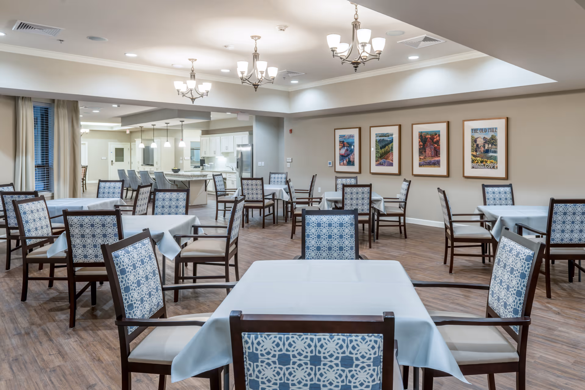 A spacious dining room in a senior living facility with multiple tables covered in white tablecloths and surrounded by wooden chairs with patterned blue and white cushions. The room features hardwood flooring, beige walls adorned with framed artwork, and modern chandeliers hanging from the ceiling. In the background, there is an open kitchen area with white cabinetry and stainless steel appliances.