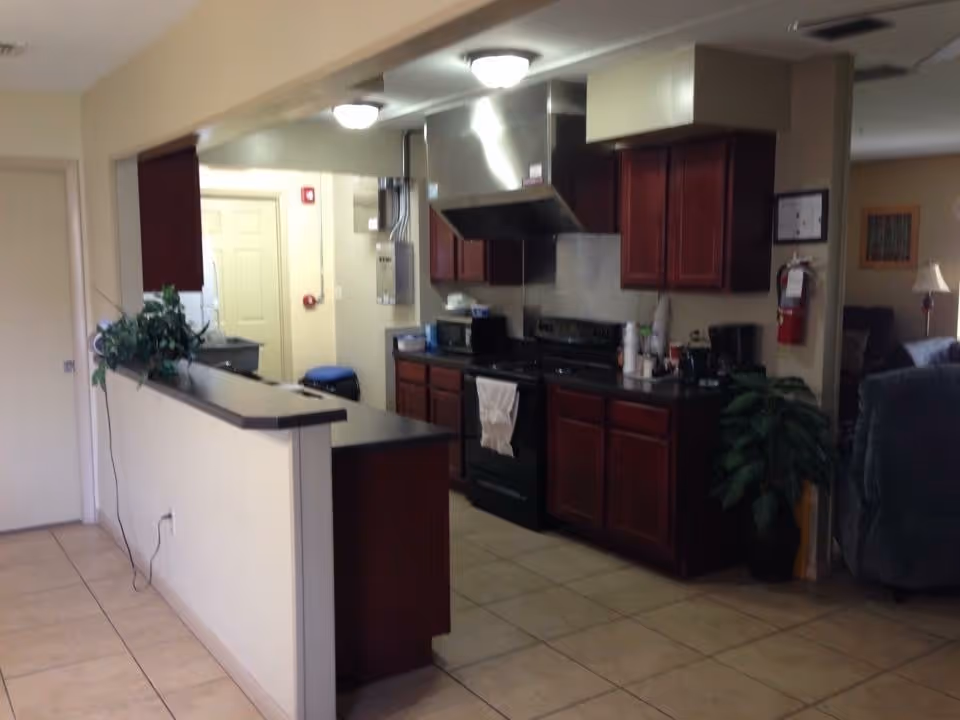 Interior view of a kitchen area in a senior living facility with dark wood cabinets, a black stove with a white towel hanging on the handle, a microwave, and various kitchen appliances on the counter. There is a half wall with a plant on it separating the kitchen from an adjacent living room area with a recliner and lamp visible.