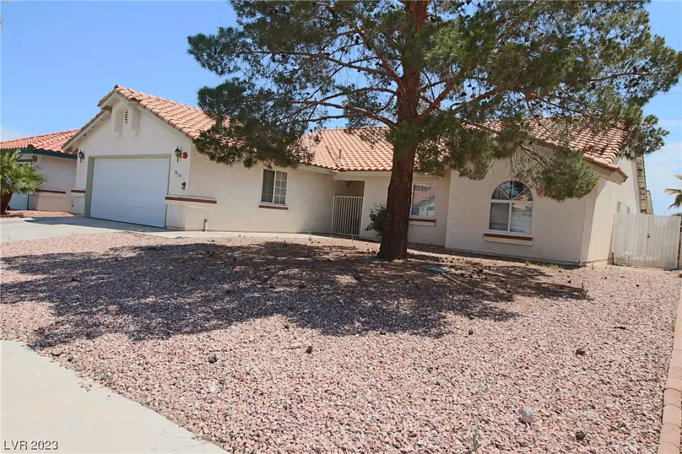 Single-story stucco house with a clay tile roof, an attached garage, a gravel front yard and a large tree casting shade.