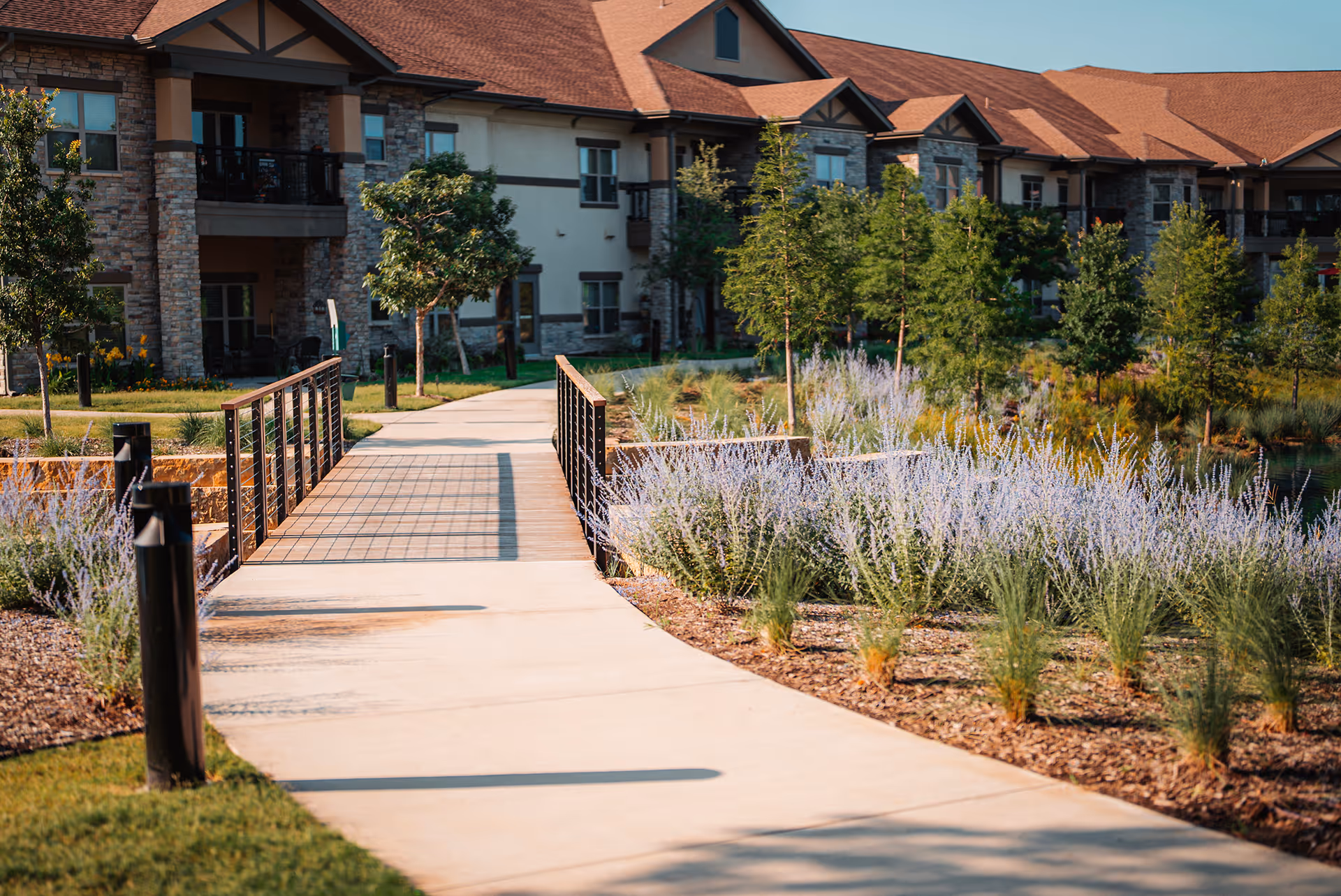 A paved walkway leading to a small bridge surrounded by landscaped gardens with purple flowering plants and young trees, in front of a large residential building with stone and beige exterior walls and a red roof under a clear blue sky.