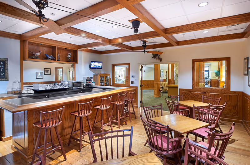 Interior view of a common area with a wooden bar counter and several high wooden bar stools. There are multiple wooden tables with matching chairs featuring red cushions. The ceiling has wooden beams and ceiling fans. A small TV is mounted on the wall above a wooden cabinet. Doorways lead to other rooms with visible seating areas.