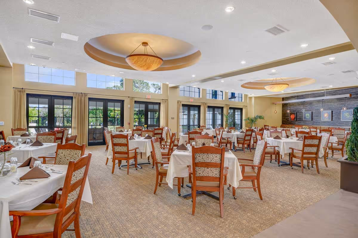 A spacious dining room in a senior living facility with multiple tables covered in white tablecloths, each set with brown napkins, glasses, and silverware. The room features large windows and glass doors letting in natural light, beige walls, carpeted floor, and decorative ceiling lights. There are wooden chairs with patterned cushions around the tables and some framed artwork on the far wall.