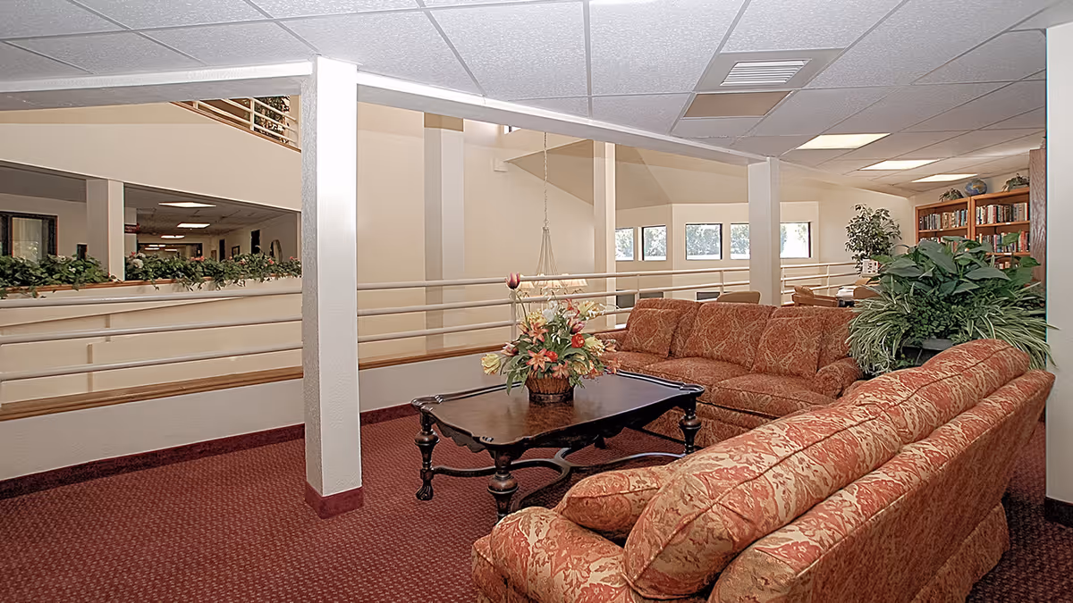 Interior view of a senior living facility lounge area with patterned red sofas arranged around a dark wooden coffee table holding a floral arrangement. The space features a red carpet, white walls, large windows, and a bookshelf with plants nearby.