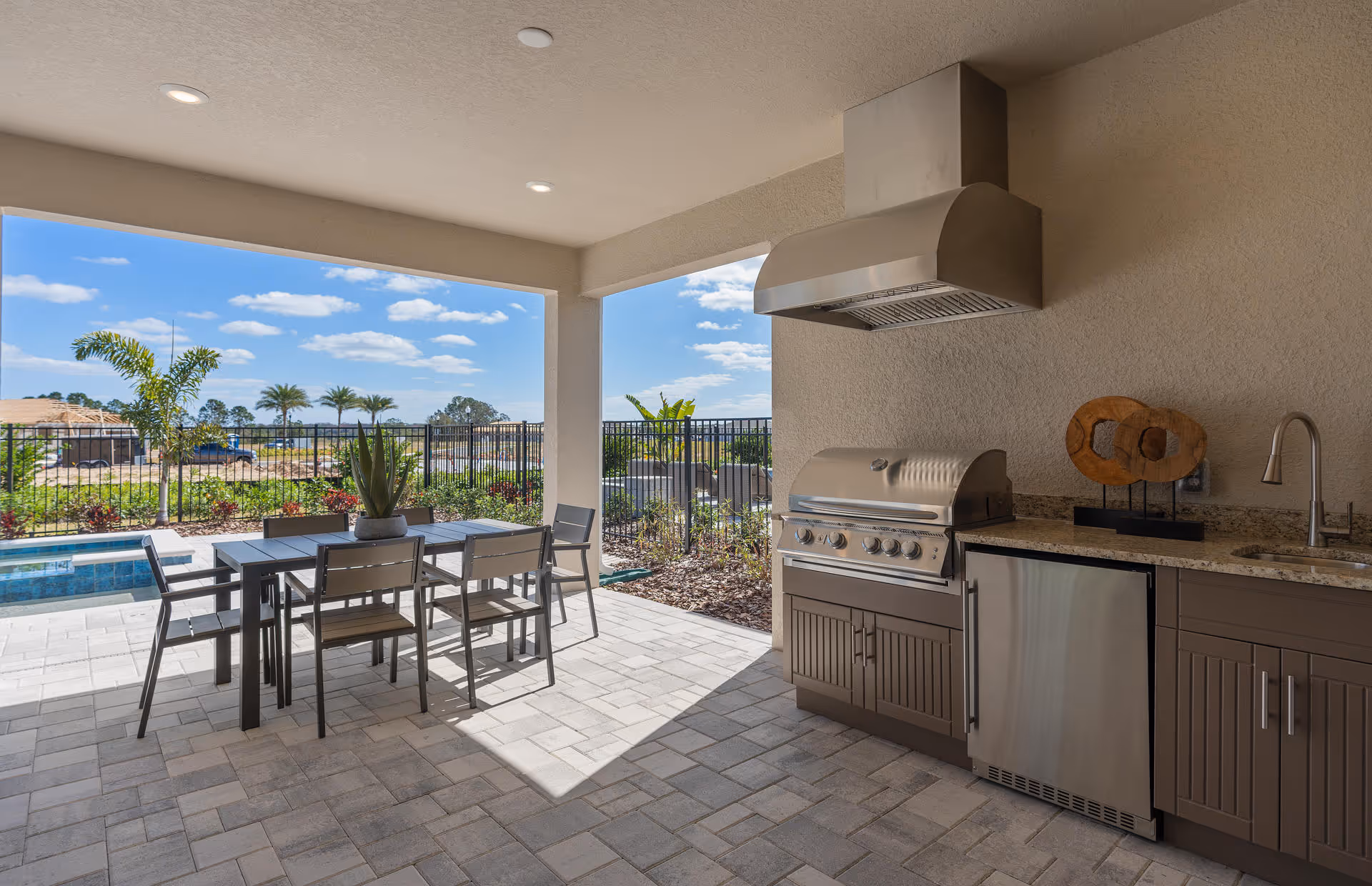 Covered outdoor patio area with a dining table and six chairs, a built-in stainless steel grill with a vent hood, a small refrigerator, a sink, and decorative wooden art on the countertop. The patio overlooks a fenced garden with palm trees and a clear blue sky with scattered clouds.