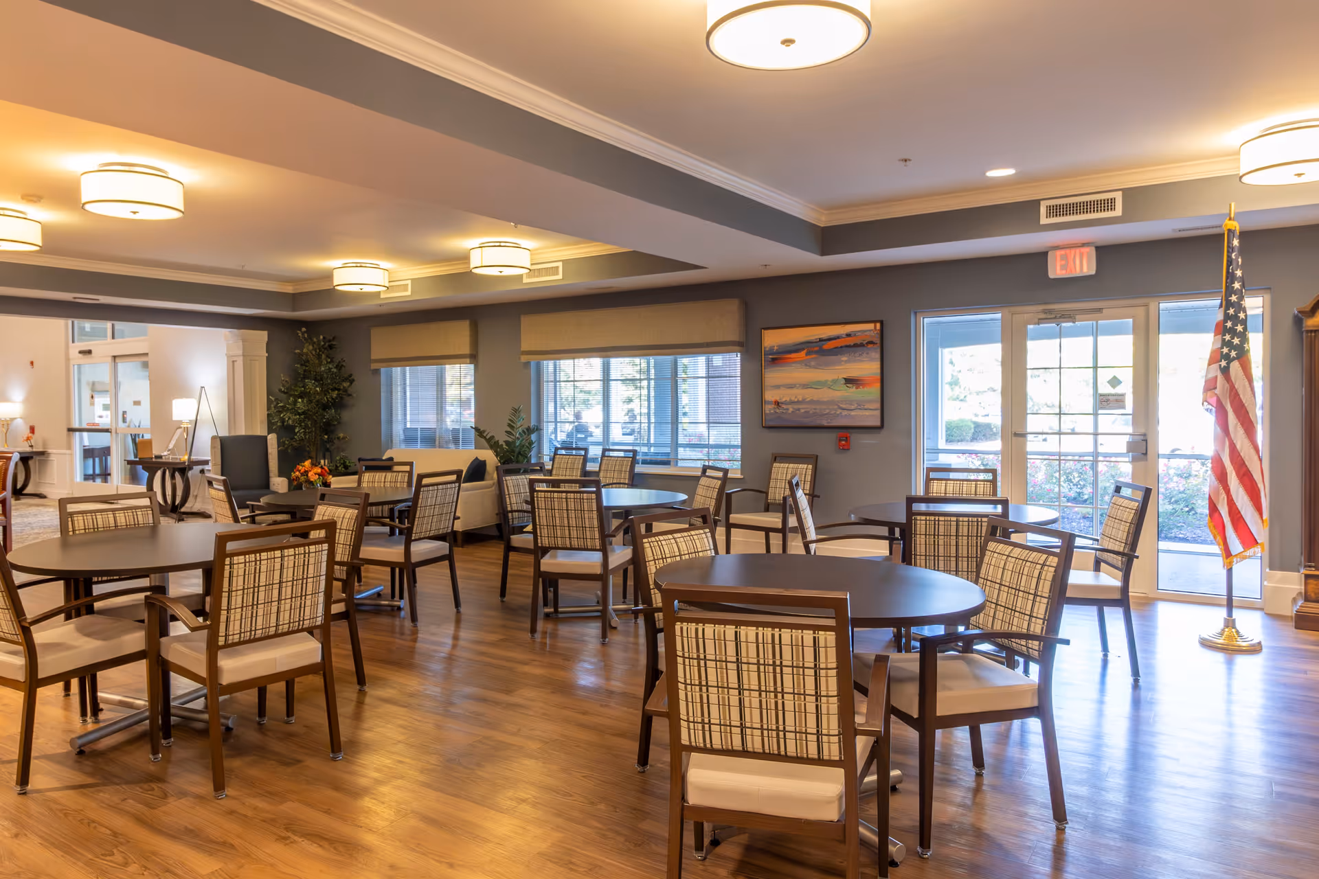 A well-lit dining room with multiple round tables and wooden chairs with plaid upholstery. The room has large windows with beige blinds, wooden flooring, ceiling lights, and an American flag near the glass exit doors. There is a painting on the wall and some plants in the corner.