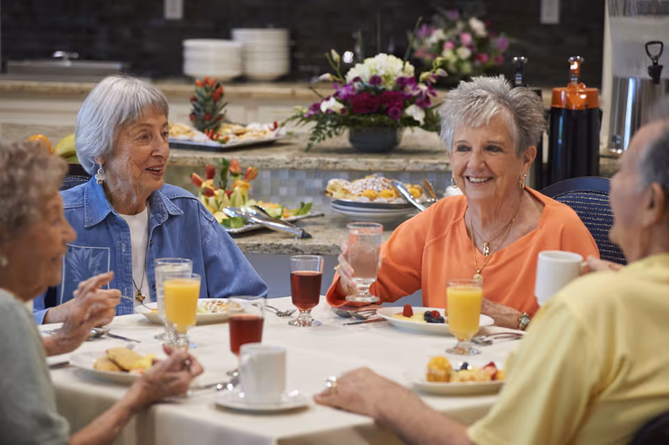 Four elderly people sitting around a dining table enjoying a meal together, with plates of food and glasses of juice and water. The background shows a buffet area with plates, flowers, and food trays.
