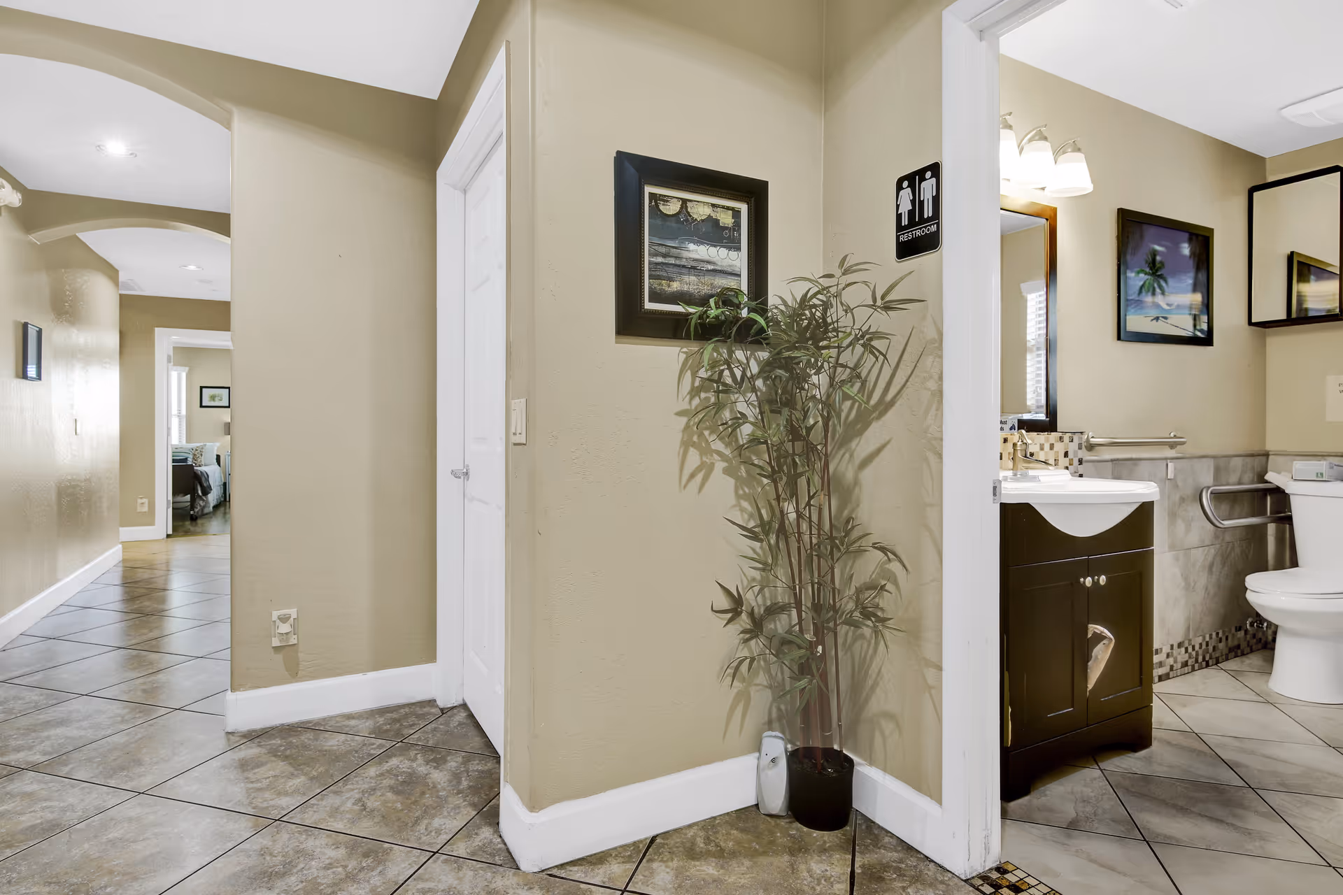 Interior view of a hallway in an assisted living facility with beige walls and tiled floors. A restroom is visible on the right side with a sink, toilet, and handrails. There is a potted plant and framed artwork on the wall near the restroom entrance. The hallway extends to a bedroom area in the background.